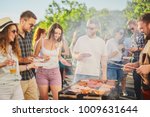 Group of people standing around grill, chatting, drinking and eating. 
