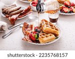 Breakfast table with waffles, croissants, sausage and bacon served maple syrup pouring over