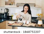 Smiling pretty Chinese woman pouring coffee in cup when eating breakfast in kitchen