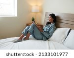 Smiling young woman in silk pajamas sitting on bed and reading ebookk on tablet computer