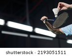 Hands of male player holding racket and hitting shuttlecock on dark badminton indoor court
