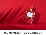 Transparent bottle of perfume with label on a red background. Fragrance presentation with daylight. Trending concept in natural materials palm leaves shadow. Women's and men's essence.