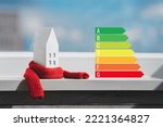 Miniature house in a red scarf and energy efficiency chart on the windowsill. The concept of passive house heating. Thermal insulation of a building or dwelling. Energy crisis.