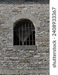 Small photo of Round arch and semicircular transom barred window with cast-iron grilles and folded white curtain cut into an Ottoman-era house stone wall in the Clock Tower area of the citadel. Gjirokaster-Albania.