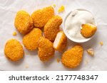 Small photo of Fried gluten free cornflake crumb chicken nuggets next to a white ceramic bowl of white sauce on white baking paper. Dipped nugget.Top view.