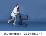 Full length portrait of young Asian woman pushing an empty shopping cart or shopping trolley isolated on deep blue background