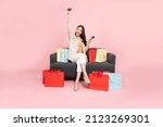 Full length portrait of Excited Asian woman sitting on sofa with shopping bags and hands up with credit card and mobile phone isolated on pink background, Shopper or shopaholic concept