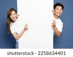Small photo of Young Asian couple is standing behind the white blank banner or empty copy space advertisement board and showing mini heart sign isolated on blue background, Looking at camera