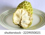 Small photo of Closeup of full ripe Indian custard apple fruit and an opened fruit in foreground showing pulpy interior and formation and arrangement of seeds isolated against plain white background.