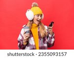 Small photo of Young beautiful woman wearing winter muffs isolated on red background holding coffee to take away and a mobile