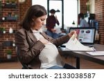 Small photo of Marketing company pregnant employee analyzing future business plan regarding profits. Expectant woman reviewing agency documentation and business analytics results while sitting at office desk.