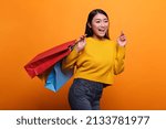 Small photo of Overjoyed excited beautiful asian woman carrying shopping bags from outlet store while wearing yellow sweater. Happy positive fashionable shopper smiling heartily while on orange background.