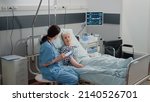 Small photo of Nurse giving assistance to senior patient with disease in bed. Medical assistant and doctor doing healthcare checkup for pensioner with oxygen tube and IV drip bag in hospital ward.