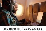 Small photo of African american passenger in airplane seat flying on work trip, using laptop on flight. Travelling with commercial airline during sunset, wearing headphones on aviation jet. Handheld shot.