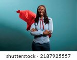 Small photo of Confident looking mighty powerful young adult person wearing superhero costume on blue background. Portrait of happy brave woman with superpowers wearing red hero cape while standing with arms