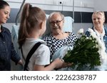 Small photo of Sick grandfather receiving flowers from granddaughter in hospital room. Bedridden old man being visited by relatives in geriatric clinic room. Little girl giving flowers to bedridden elderly man.