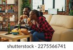 Small photo of Cheerful young people eating hamburgers and fries in living room, watching favorite film on tv and drinking alcohol. Boyfriend and girlfriend serving fast food order from takeout place.