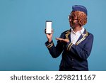 Small photo of Female flight attendant wearing uniform holds cell phone with empty screen for advertising isolated on blue background. Professional african american stewardess looking at camera in studio shot.