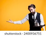 Small photo of Friendly gracious waiter makes way for people to enter restaurant, providing great service and amazing food at five star place. Young adult with suit and tie posing against yellow background.