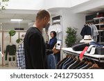 Small photo of Male customer grasping cellphone that shows website of a boutique. Caucasian client shopping near clothes racks while holding mobile device displaying discounted online clothing prices.