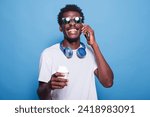 Small photo of Portrait of African American guy with sunglasses talking on mobile device while holding a beverage. Black man with wireless headphones around neck is laughing on phone call and grasping a coffee cup.