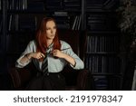 Small photo of A young red-haired witch with a magic wand and a haughty expression sitting in an old library, witchcraft books and old clock on the table. She is dressed in a white shirt, black skirt and green tie