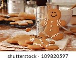 Smiling gingerbread man standing next to flour sifter with baking ingredients and additional gingerbread cookies in background.  Partially decorated cookies in foreground.  Closeup with shallow dof.