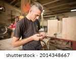 Small photo of Carpenter artisan resting in workshop and scrolling on smartphone. Handsome handicraftsman texting message and tapping on phone