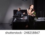 Small photo of Disdainful attractive young woman sitting on the bed dressed in elegant black evening wear ignoring her husband or boyfriend as he pleads with her to forgive him or overlook an indiscretion