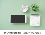 white smartphone, included clipping path on touchscreen,  white analog alarm clock, calendar, small green tree on green background for workspace concept