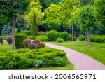 curved pedestrian walkway of stone tiles in park with landscape design with green plants bushes and trees, landscaped of thujas and deciduous buhes on the lawn, nobody.