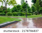 Small photo of Irrigation system landscaping watering in park with plants, water spray moistening in backyard with tile pavement and curb, iron lattice drainage system and pillar lantern for garden lighting, nobody.