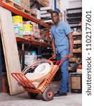 Small photo of Focused African American diligent positive carrying handbarrow with construction supplies purchased in shop of building materials