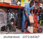 Small photo of Young African-American man putting materials for overhauls on handbarrow in building materials store