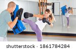 Small photo of Concentrated woman practicing self-defense techniques in gym, kicking boxing shield in hands of her coach