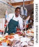 Small photo of Friendly man seller proffering fresh squid in fish marke