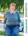 Positive mature woman with handbag sitting on bench in city park and smiling.