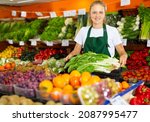Small photo of Portrait of a positive girl working part-time as a trainee seller in the store, standing in the vegetable department, ..holding a crate of precocious Chinese cabbage