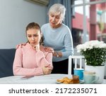 Small photo of Senior woman calming chagrined young adult daughter sitting at table at home .