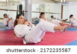 Sporty young woman in white kimono warming up before group martial arts training in gym, doing abdominal crunch..