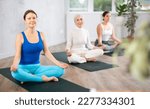 Small photo of Sedulous women doing lotus pose of yoga on black mat in light gym room with pot plants