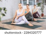 Small photo of Sedulous women doing spinal twist pose of yoga on black mat in light gym room with pot plants