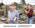 Small photo of Two upset young and old female neighbors disagree with each other while digging garden on smallholding in autumn