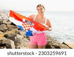 Young smiling woman in summer clothes posing with big flag of Croatia on sea coast