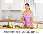 Small photo of Portrait of positive girl in nightdress standing at kitchen table at home, slicing vegetables, cooking salad.