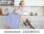 Small photo of Housewife works in cozy kitchen. Woman holds mobile phone in one hand and communicates with friend, in other hand she holds vacuum cleaner handle and furbish carpet near dining table