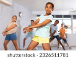 Small photo of African girlie performs movements during warm-up, limbering-up part of workout together with peers. Group of young girls and guys dance modern waacking in fitness club unfocused