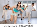 Small photo of Positive female teenager dances shuffle in choreographic school, group of young people in sportswear and dark glasses ball cap trains together with their classmates in gym before competitions, battle