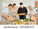 Small photo of Amiable young guy in black chef jacket and hat, running culinary courses for mixed age group of ordinary people, sharing secrets of cooking salmon