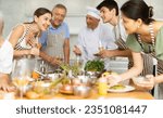 Small photo of Positive friendly woman participating in group culinary masterclass led by professional chef, enjoying wine, engaging in cheerful conversations with other participants, and cooking together at table
